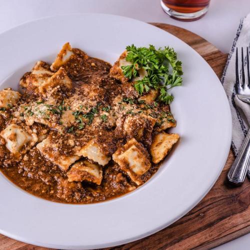 A plate of ravioli topped with meat sauce, herbs, and grated cheese, served on a wooden board with a fork and knife.