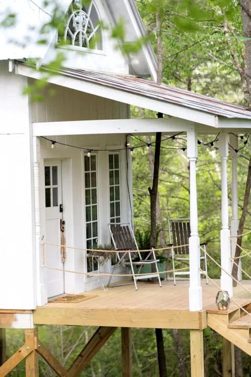 A charming treehouse with a porch, two chairs, and string lights, surrounded by lush greenery.