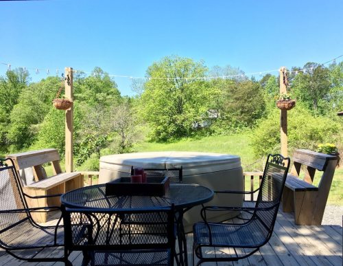 A sunny deck with a hot tub, surrounded by green trees, featuring a table and chairs for outdoor relaxation.