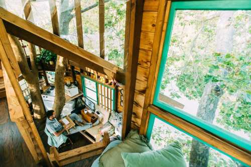 A cozy treehouse interior with a person working at a desk surrounded by large windows and greenery.