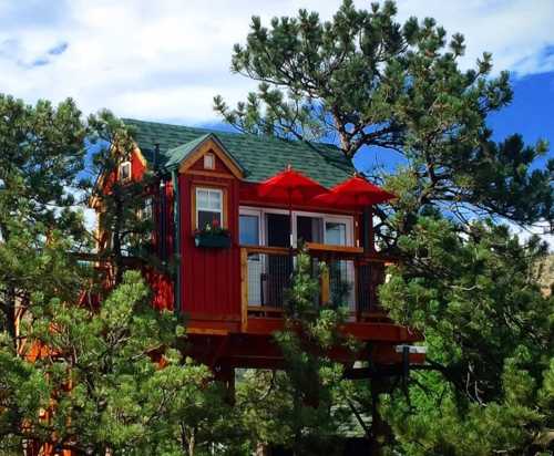 A colorful treehouse nestled among green trees, featuring a balcony and red awnings under a blue sky.