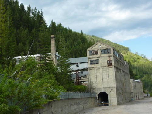 Abandoned industrial building surrounded by dense forest and mountains under a cloudy sky.