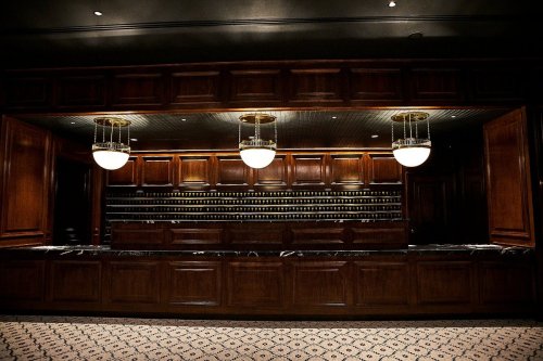 A dimly lit bar with wooden cabinetry, marble countertops, and elegant pendant lights, featuring shelves of bottles in the background.