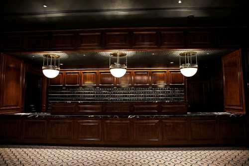 A dimly lit bar with wooden cabinetry, marble countertops, and elegant pendant lights, featuring shelves of bottles in the background.