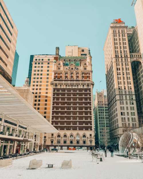 A snow-covered urban plaza surrounded by tall buildings, featuring a historic brick building with ornate architecture.