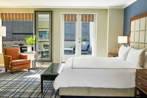 A modern hotel room featuring a bed, armchair, mirror, and balcony access with natural light.