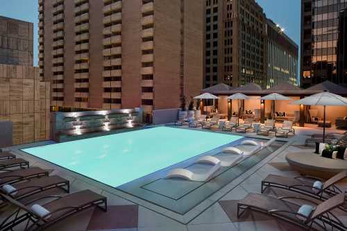 Rooftop pool area with lounge chairs and umbrellas, surrounded by tall buildings at dusk.