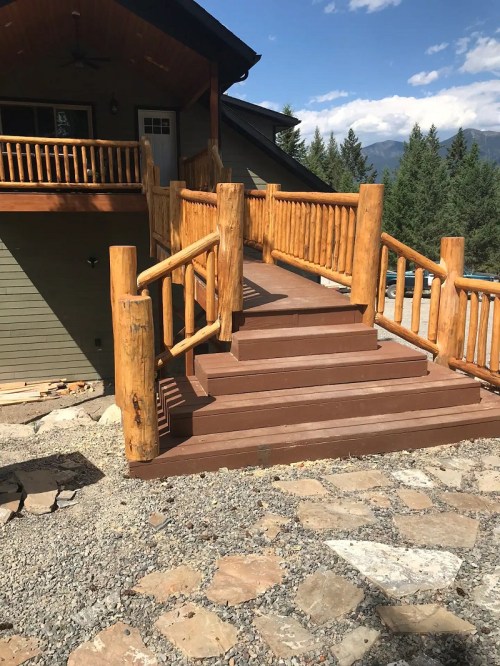 Wooden staircase with log railings leading up to a house, surrounded by trees and a clear blue sky.