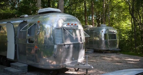 Two shiny Airstream trailers parked in a wooded area, surrounded by trees and gravel.