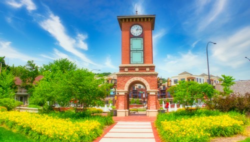 A brick clock tower surrounded by vibrant greenery and yellow flowers under a blue sky with wispy clouds.