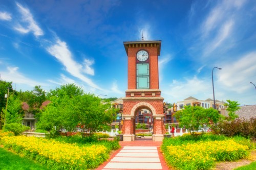A brick clock tower surrounded by vibrant greenery and colorful flowers under a blue sky with wispy clouds.