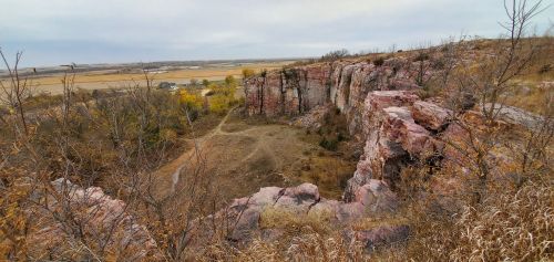 A panoramic view of rocky cliffs with sparse vegetation, overlooking a valley and distant fields under a cloudy sky.