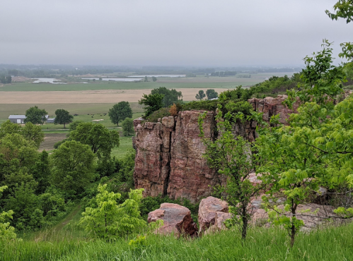 A rocky cliff overlooks a lush green valley and fields under a cloudy sky.