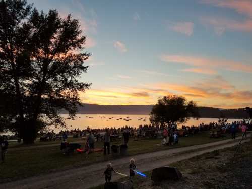 A sunset over a lake with a crowd gathered on the shore, two children playing with light-up toys in the foreground.