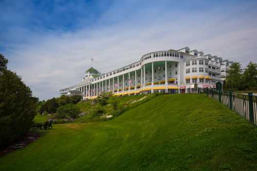 A large, white hotel with a green roof sits on a grassy hill under a cloudy sky, surrounded by trees and flags.