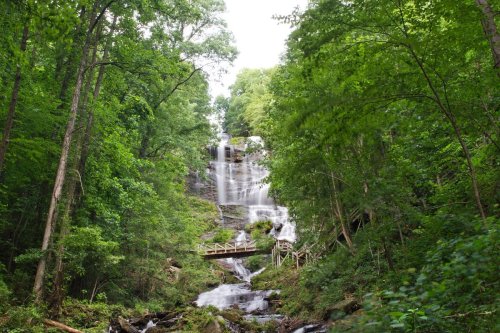 A cascading waterfall surrounded by lush green trees, with a wooden bridge crossing over a stream below.