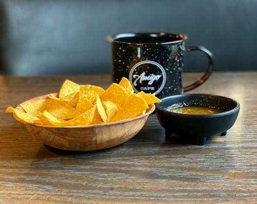 A wooden bowl of tortilla chips next to a small black bowl of salsa and a black mug on a wooden table.
