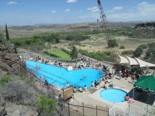 Aerial view of a crowded pool area with people swimming and lounging, surrounded by a scenic landscape.
