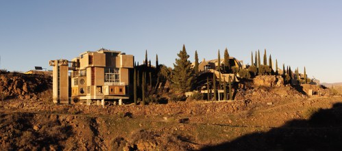 A modern building on a rocky hillside, surrounded by tall trees and a clear blue sky at sunset.