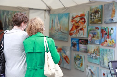 Two women admire colorful paintings displayed in a tent at an art event.