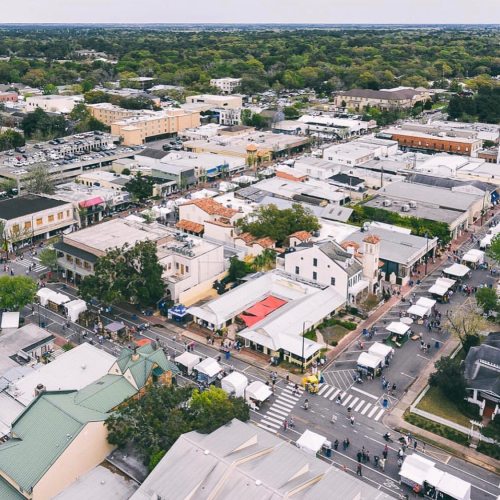 Aerial view of a bustling market street with tents, shops, and greenery in a vibrant town.