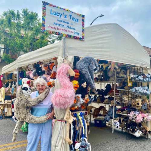 An elderly woman stands in front of a toy stall, holding a sloth and surrounded by stuffed animals at a market.