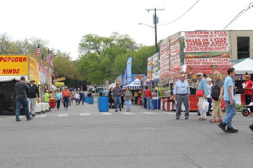 A bustling street fair with food stalls, colorful banners, and people walking along the street.
