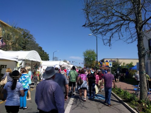 A bustling street fair with people walking among vendor tents under a clear blue sky.