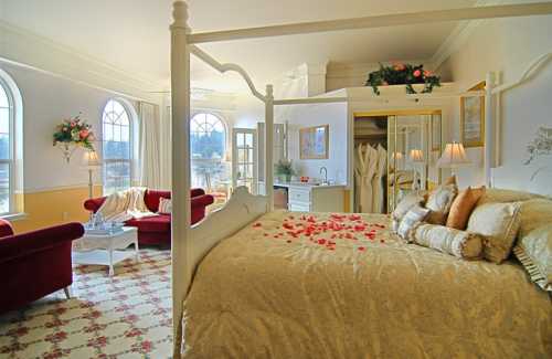 A cozy hotel room featuring a four-poster bed, rose petals, a seating area, and large windows with scenic views.