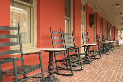 A row of black rocking chairs with a small table on a brick porch, against a red wall and large windows.