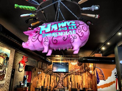 A large pink pig sculpture hangs from the ceiling in a smokehouse restaurant, with colorful tools and decor around it.