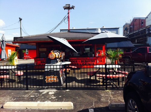 A colorful outdoor eatery with picnic tables, umbrellas, and a BBQ grill, surrounded by a black fence and industrial buildings.