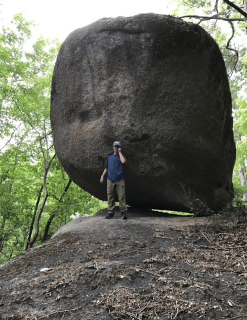 A person stands beside a large, rounded boulder on a rocky surface, surrounded by green trees.