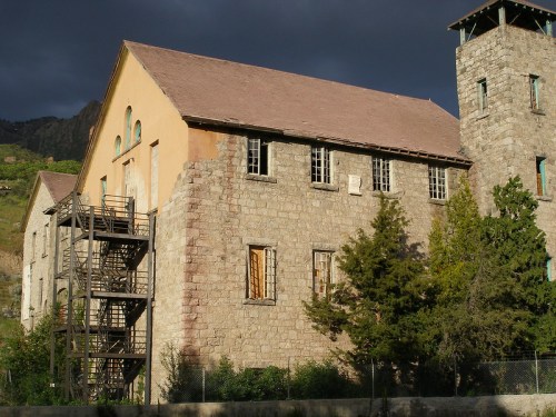 Abandoned stone building with a fire escape, surrounded by trees and mountains under a cloudy sky.