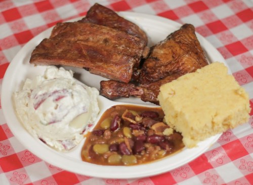 A plate of barbecue ribs, potato salad, baked beans, and cornbread on a red and white checkered tablecloth.