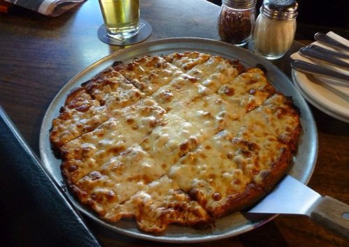 A freshly baked cheese pizza on a metal tray, served with condiments and a glass of beer in the background.