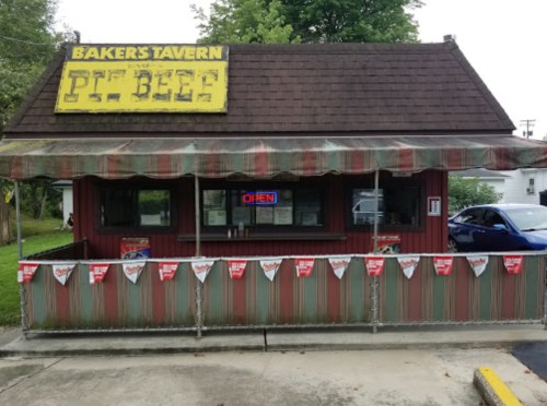 A small, rustic tavern with a yellow sign reading "Baker's Tavern" and a red "OPEN" sign, decorated with banners.