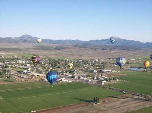 Aerial view of colorful hot air balloons over a small town and green fields, with mountains in the background.