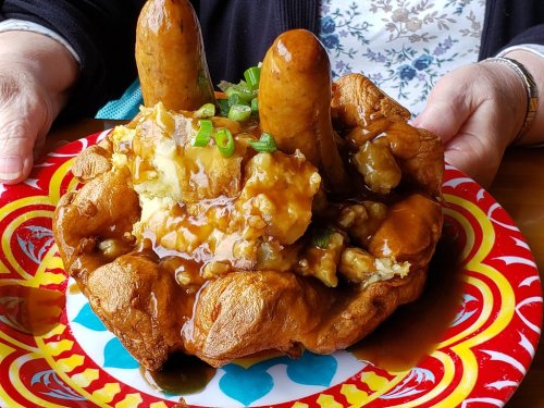 A person holds a colorful plate with a large, fluffy pastry topped with mashed potatoes, sausage, and gravy.