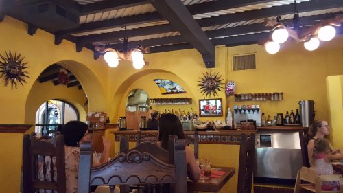 Interior of a vibrant restaurant with yellow walls, wooden beams, and decorative elements, featuring patrons at tables.