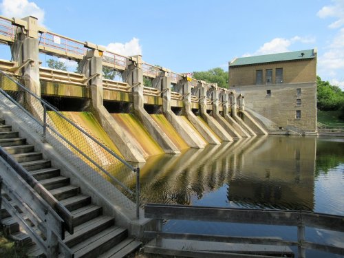 A dam with multiple spillways, reflecting in calm water, surrounded by greenery and a blue sky.