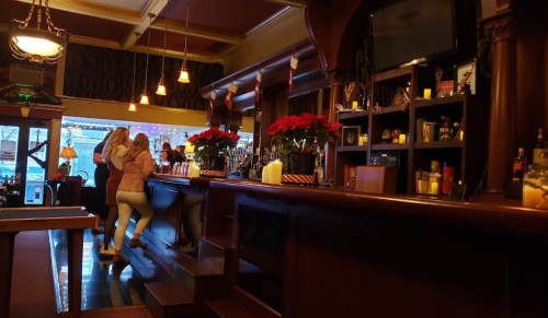 A cozy bar scene with two women chatting at the counter, decorated with holiday flowers and warm lighting.