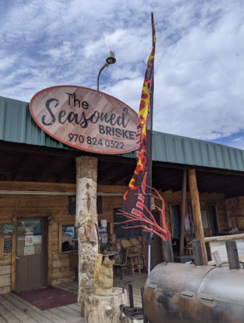 Sign for "The Seasoned Brisket" restaurant with a flag and wooden exterior in a rustic setting.