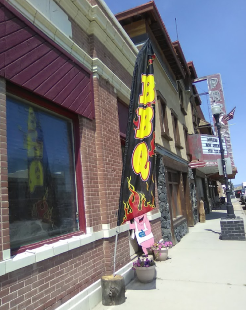 A tall BBQ flag stands outside a brick building, with flower pots nearby and a clear blue sky above.