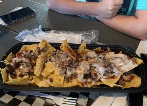 A tray of nachos topped with meat and creamy sauce, placed on a table next to a person's arm and a phone.