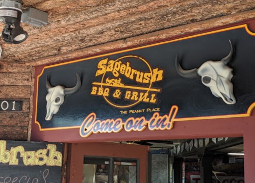 Sign for "Sagebrush BBQ & Grill" featuring bull skulls and the phrase "Come on in!" on a rustic wooden background.