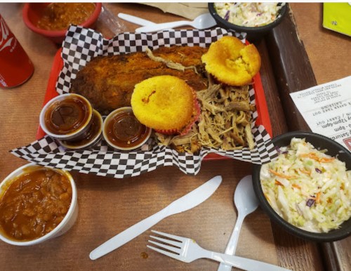 A plate of barbecue with pulled pork, cornbread muffins, coleslaw, baked beans, and dipping sauces on a checkered tray.