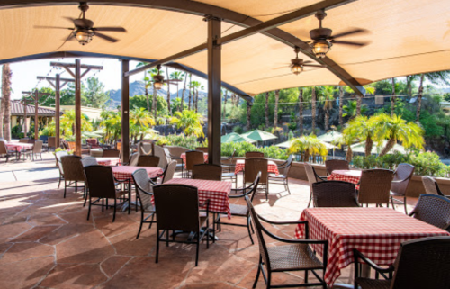Outdoor dining area with checkered tablecloths, fans, and palm trees, under a shaded patio.