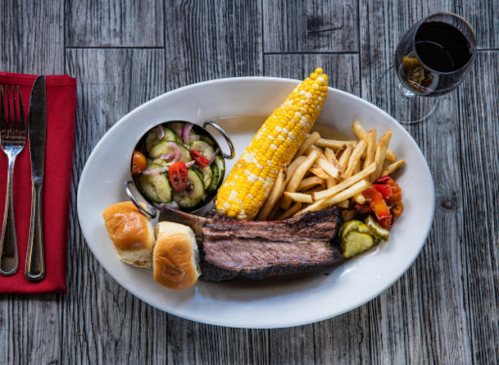 A plate with brisket, corn on the cob, fries, salad, and two rolls, accompanied by a glass of red wine.
