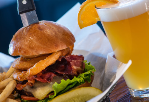 A juicy burger topped with bacon and onion rings, served with fries and a glass of beer with an orange slice.